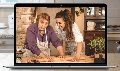 A photo of a computer showing an older lady helping a young lady make pasta