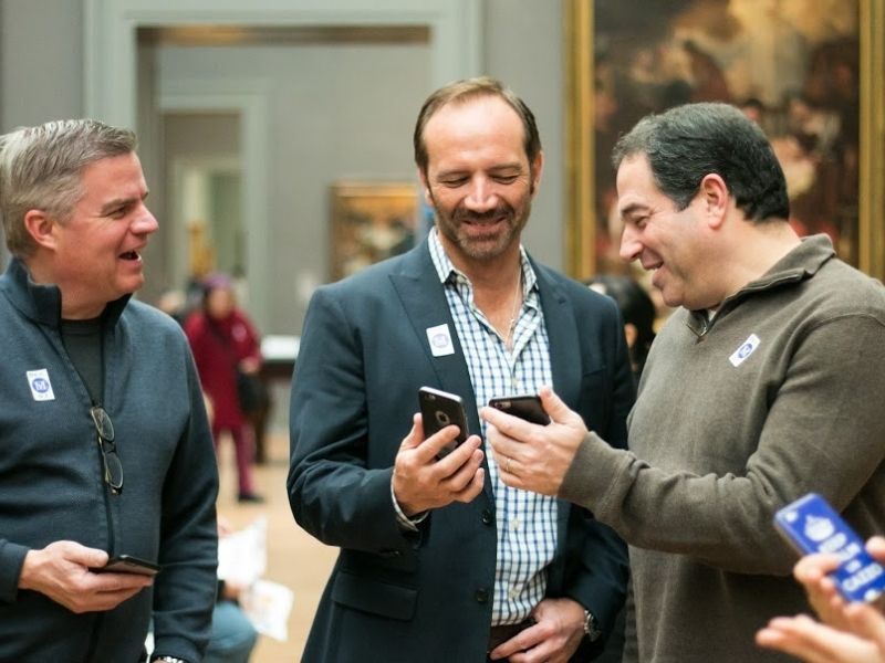 Three men gesturing to smrat phones and smiling in a museum